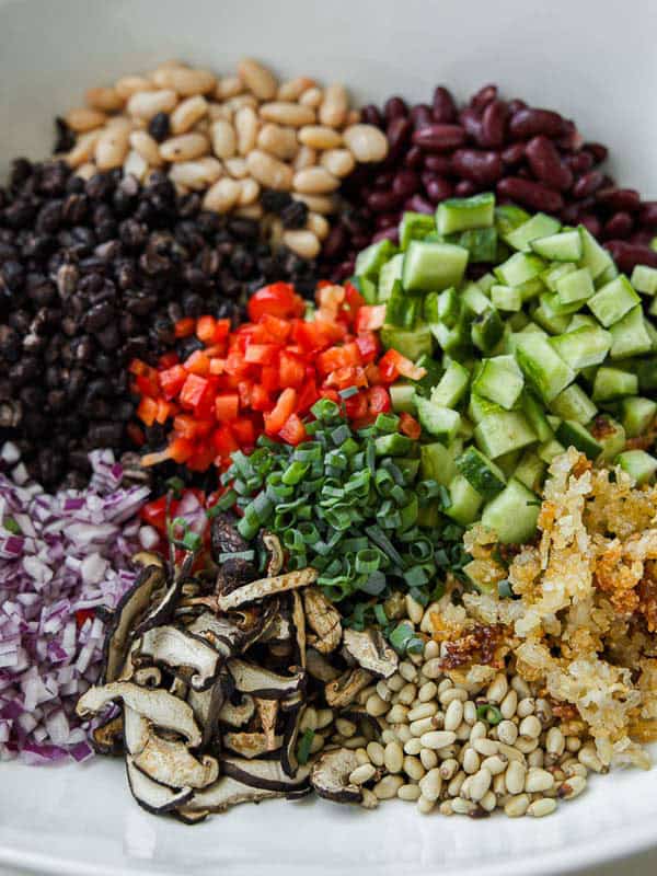 A bowl filled with chopped vegetables and beans, including cucumber, red bell pepper, red onion, black beans, kidney beans, white beans, pine nuts, green onions, sautéed mushrooms, and crispy cooked rice.